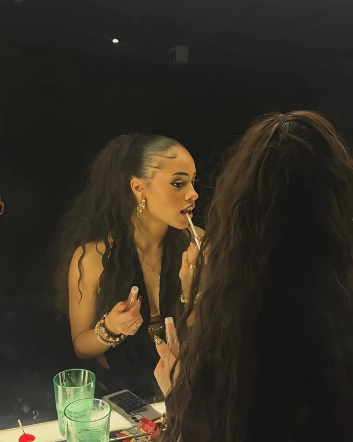 Alexia trägt abends vor einem Spiegel Lipgloss auf, das Haar in langen Wellen, goldener Schmuck.