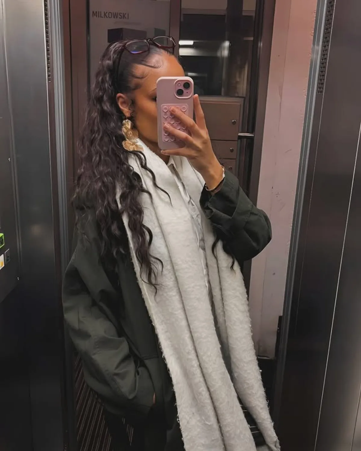 Spiegelselfie in einem Aufzug: Alexia mit langem Wellen-Zopf, oversized dunkelgrüner Jacke über hellem Hemd.